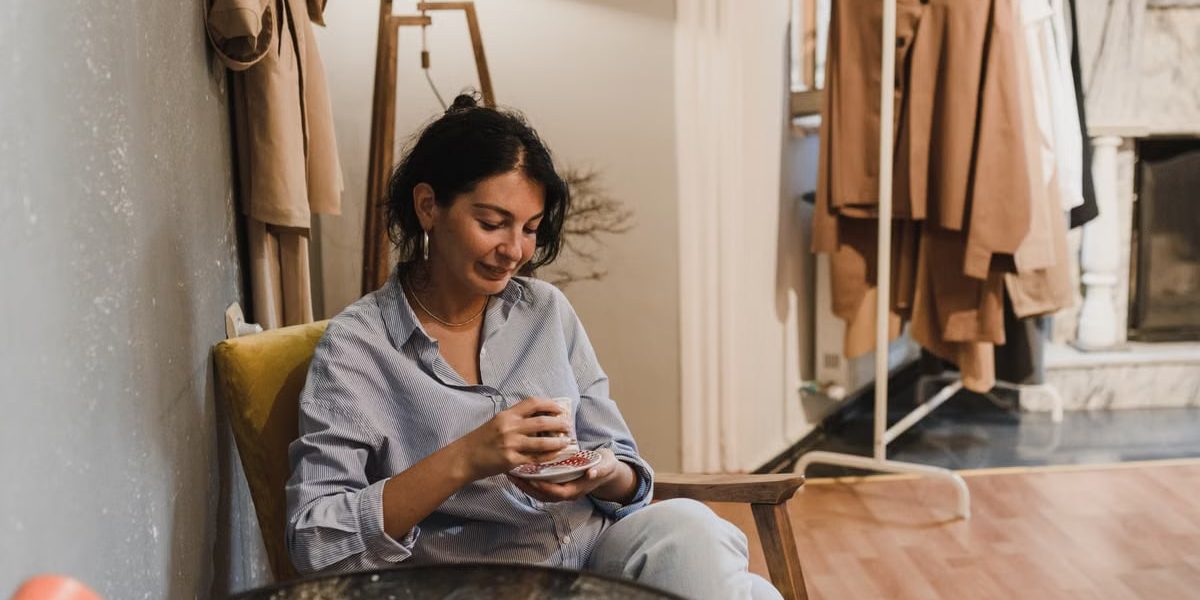 Woman sitting in a boutique, enjoying a coffee with clothes displayed nearby.