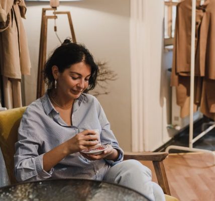 Woman sitting in a boutique, enjoying a coffee with clothes displayed nearby.