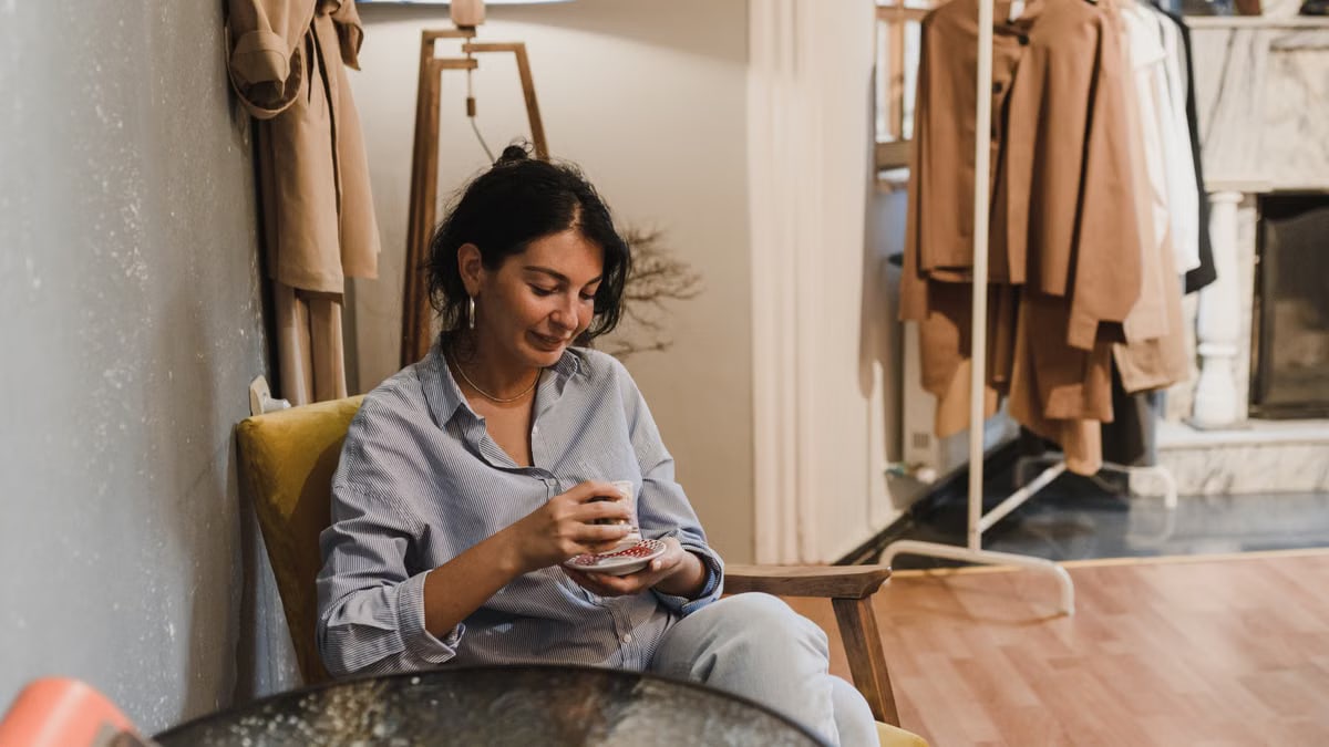 Woman sitting in a boutique, enjoying a coffee with clothes displayed nearby.