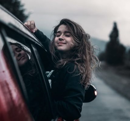 Contented young female with brown hair fluttering in wind leaning out of window of moving car in countryside