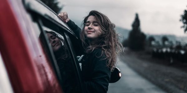 Contented young female with brown hair fluttering in wind leaning out of window of moving car in countryside